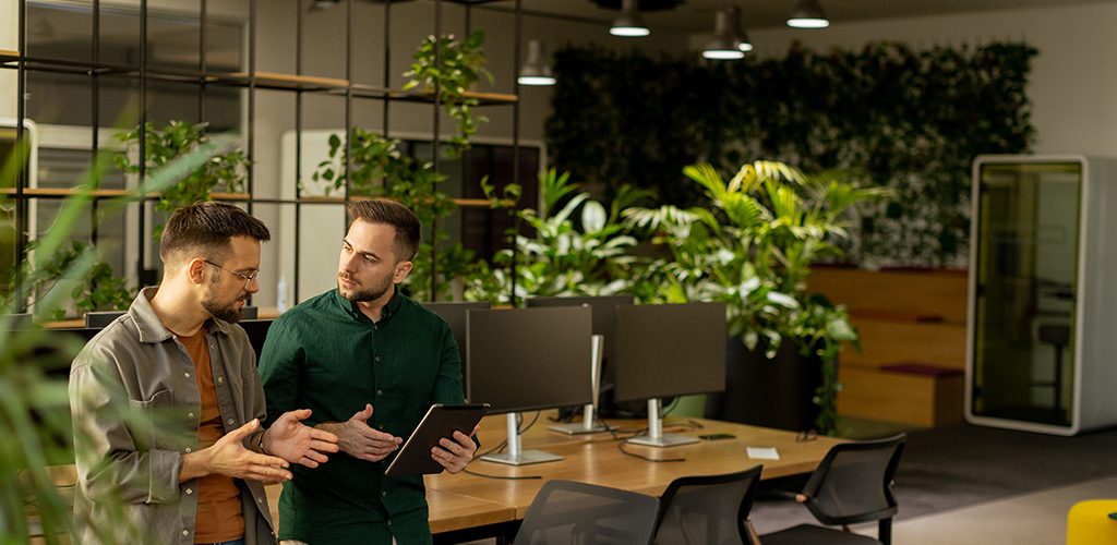 Two professionals are engaged in a focused conversation while holding digital tablet in a contemporary office space, featuring rustic exposed brickwork and warm natural light